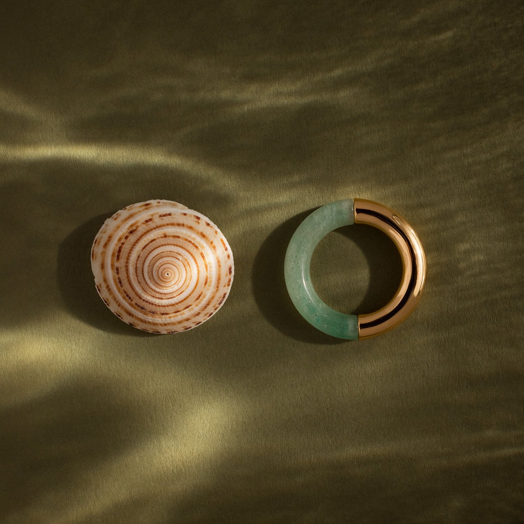 A Half Jade Ring with gold accents rests on a textured olive-green surface beside a round seashell—statement jewelry perfect for any occasion.