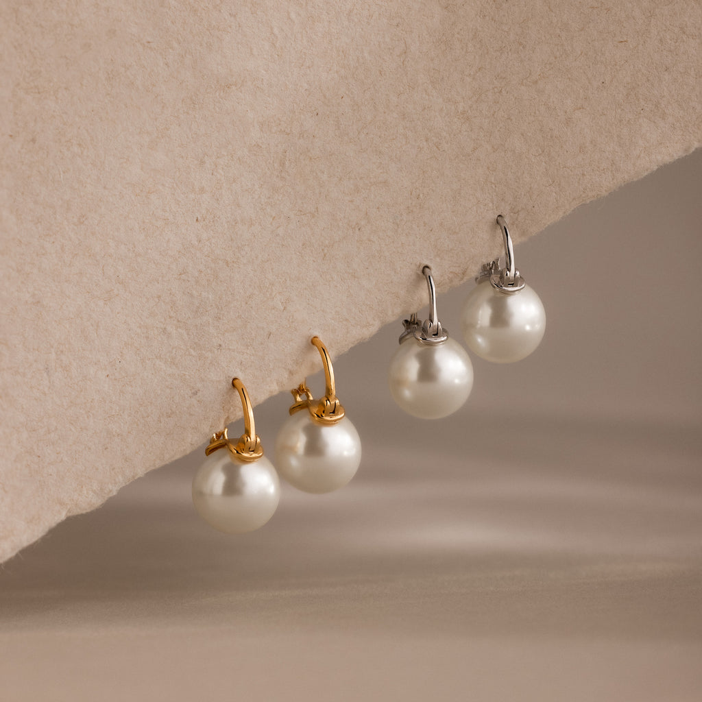Two pairs of Pearl Dome Drop Earrings, each featuring lustrous pearls—one with gold hooks and one with silver—are displayed on a textured surface. Perfect for any occasion.