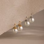 Two pairs of Pearl Dome Drop Earrings, each featuring lustrous pearls—one with gold hooks and one with silver—are displayed on a textured surface. Perfect for any occasion.
