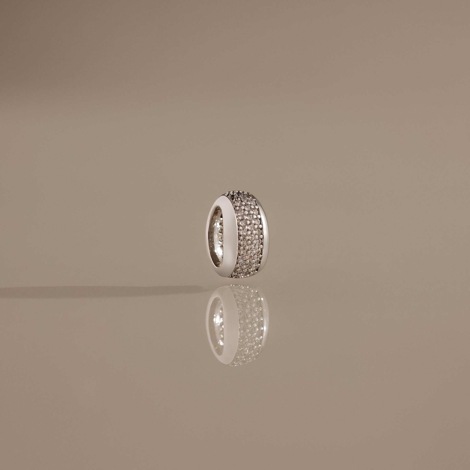 The Bold Pave Dome Charm in Silver, adorned with rows of sparkling stones and standing upright on a reflective beige surface, showcases timeless elegance in every detail.