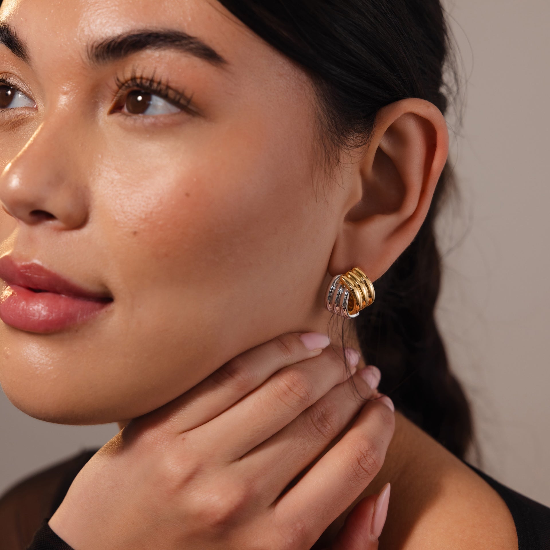 A woman with glossy skin wears Ribbed Mixed Metal Studs in gold and silver, touching her neck and gazing to the side.