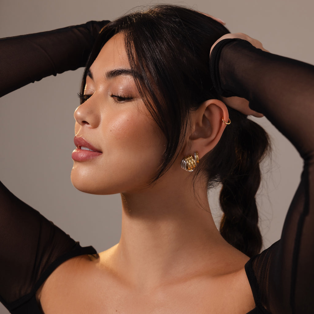 A woman with closed eyes and hands in her hair wears Ribbed Mixed Metal Studs and a sheer black top against a neutral background.
