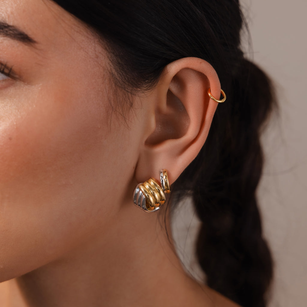 Close-up of a woman's ear adorned with Ribbed Mixed Metal Studs—sculpted ridged earrings in gold and silver for a modern look—plus a gold hoop cuff, her dark braided hair visible.