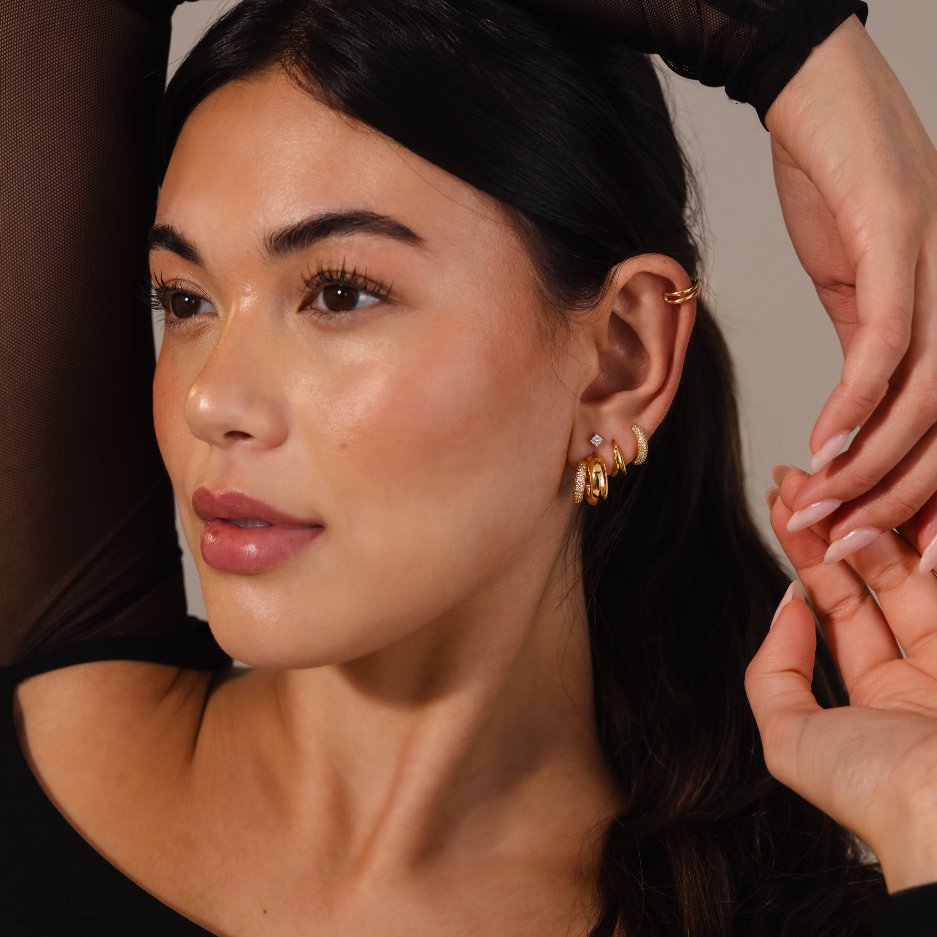 A woman with long dark hair, wearing Duo Pave Dome Studs and a cuff, poses in a black top with one arm raised—showcasing how these earrings elevate her everyday style.