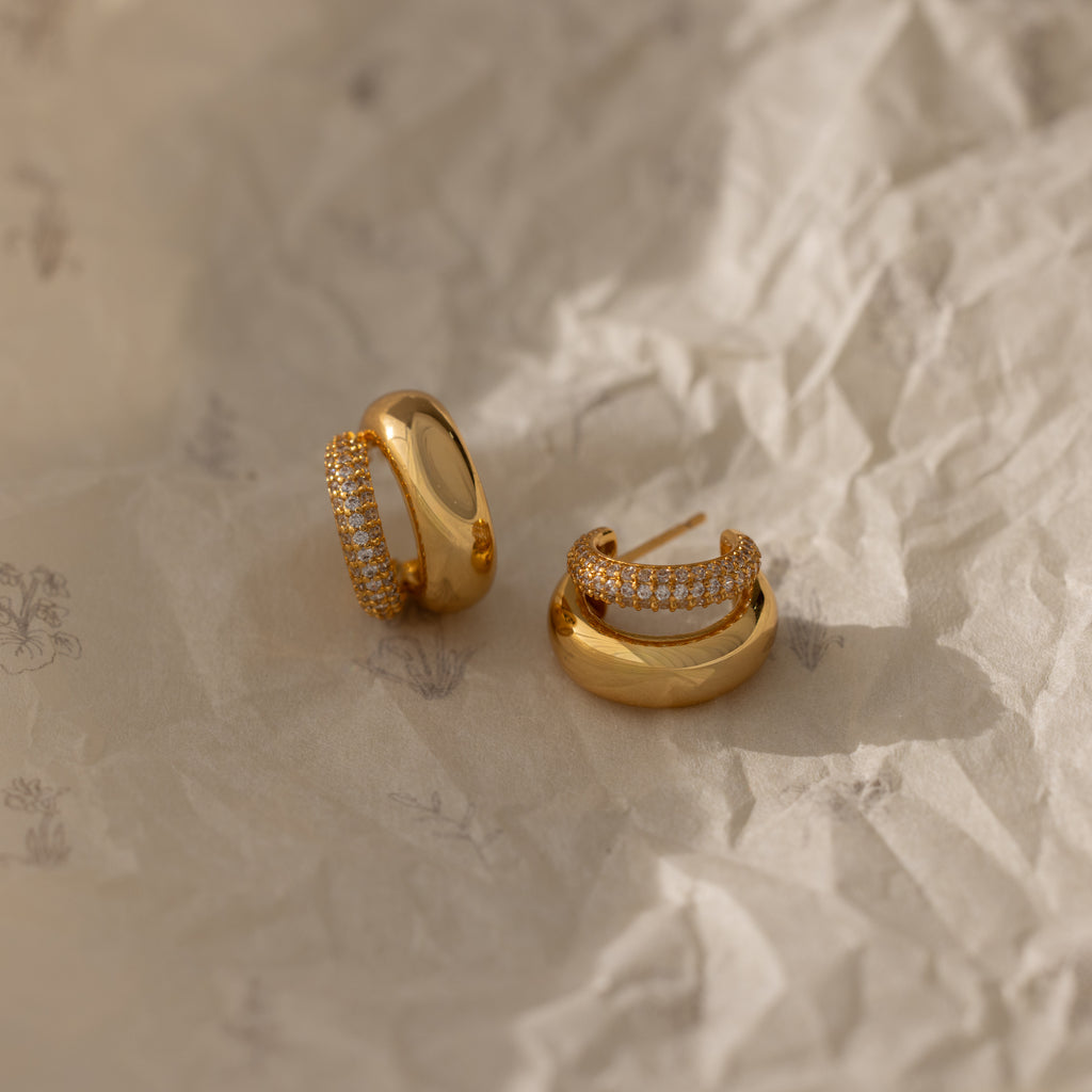 The Duo Pave Dome Studs—one plain, one with pavé diamonds—sit on crumpled beige paper, offering a perfect pair of everyday earrings inspired by classic curves.