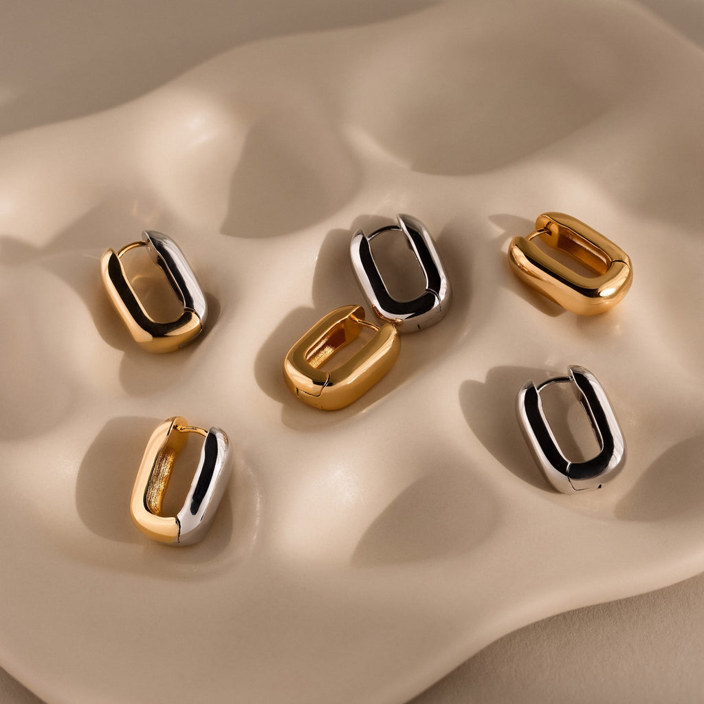 The Bold Link Hoops, along with four other gold and silver oval hoop earrings—some chunky—are displayed on a wavy, beige ceramic dish.