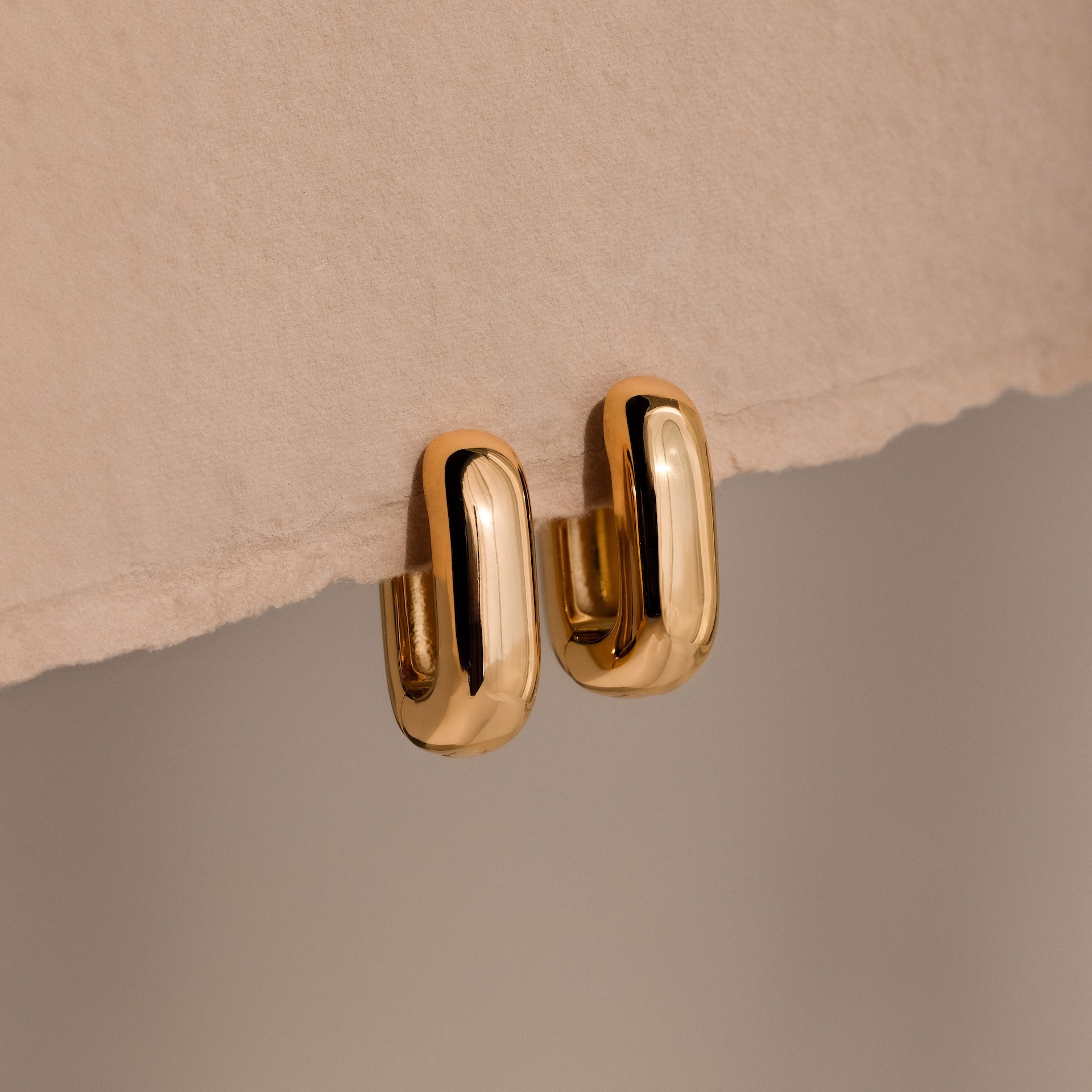 Bold Link Hoops in 18K Gold rest on a textured beige surface, showcasing a modern earring style that's perfect for everyday wear.