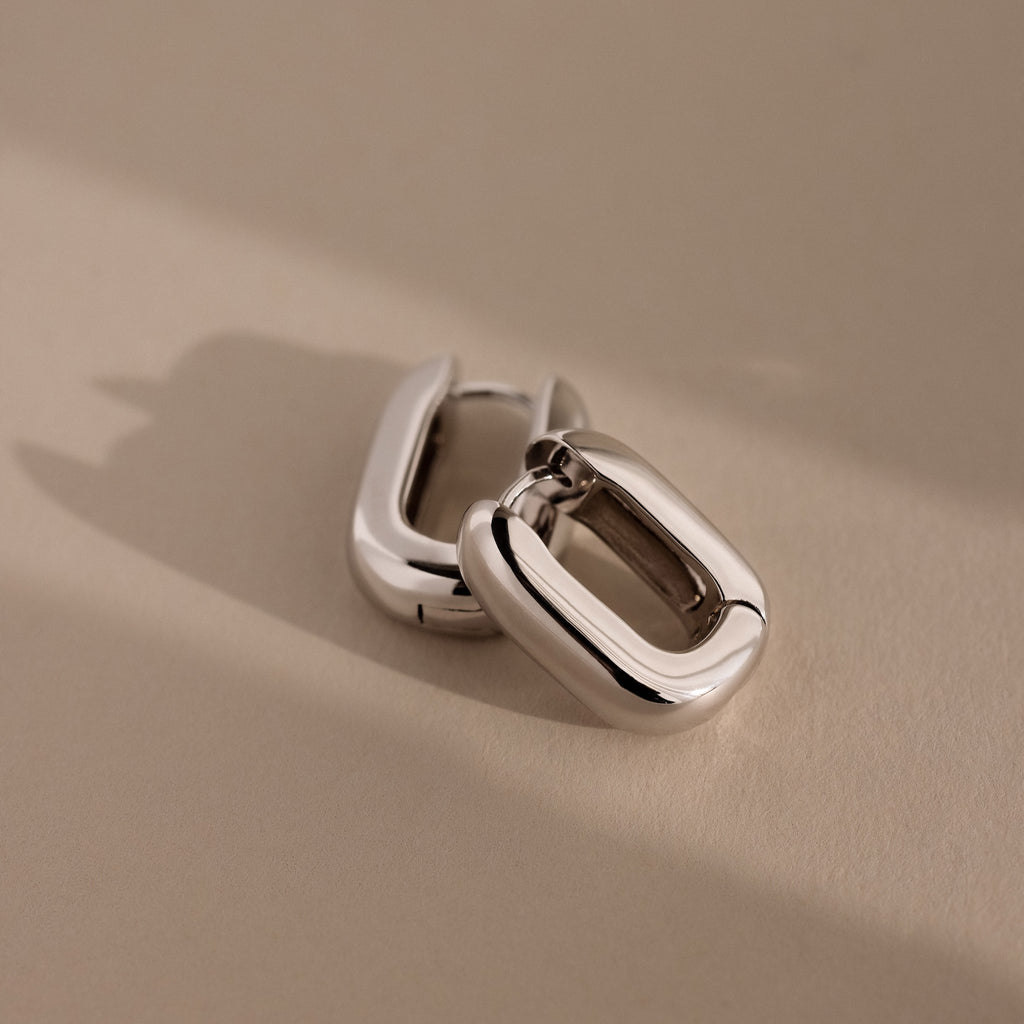 A pair of chunky, rectangular Bold Link Hoops in White Gold rests on a neutral beige surface, creating a modern jewelry statement.