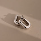 A pair of chunky, rectangular Bold Link Hoops in White Gold rests on a neutral beige surface, creating a modern jewelry statement.