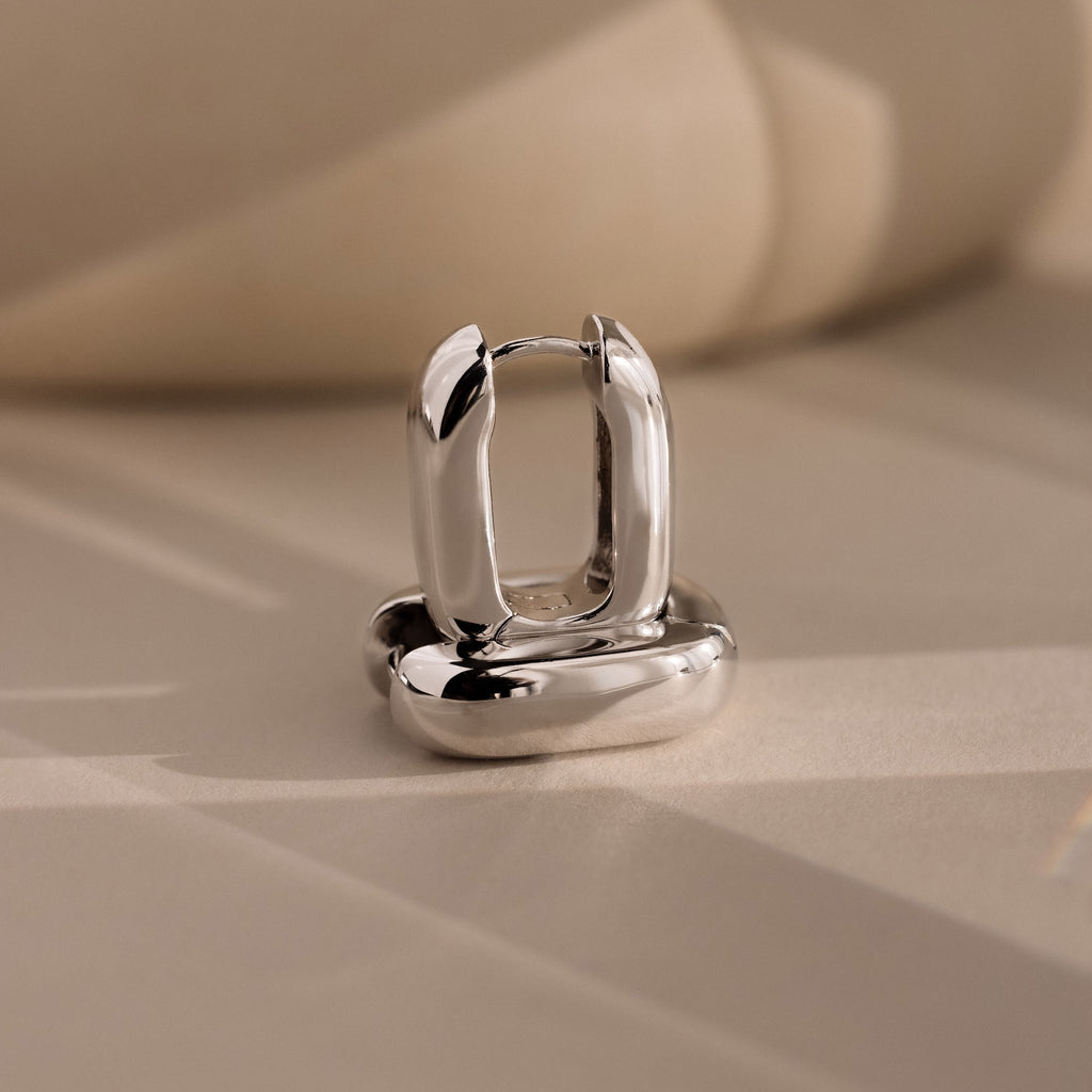 The Bold Link Hoops in White Gold, from our signature collection, stand upright on a beige surface with soft shadows—perfect for fans of modern jewelry.