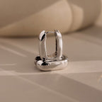 The Bold Link Hoops in White Gold, from our signature collection, stand upright on a beige surface with soft shadows—perfect for fans of modern jewelry.