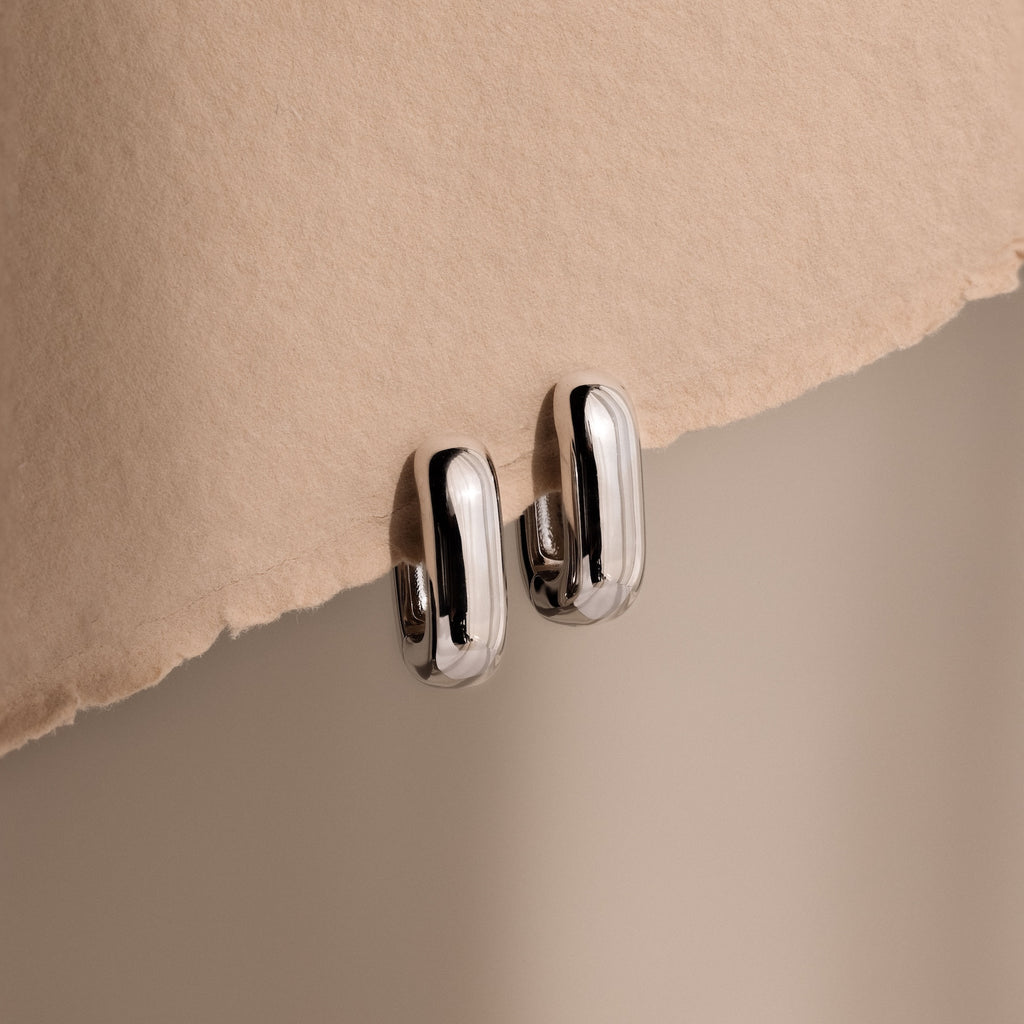 Bold Link Hoops in White Gold rest on a textured beige surface under soft lighting, highlighting the modern elegance of this jewelry piece.