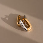 Bold Link Hoops in Mixed Metal—chunky gold and silver rectangular hoop earrings—rest on a beige surface with soft shadows, bringing a modern, stylish edge to any look.