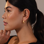 Woman with a ponytail wears Bold Link Hoops in 18K Gold, gazing left with her hand near her chin against a neutral background.
