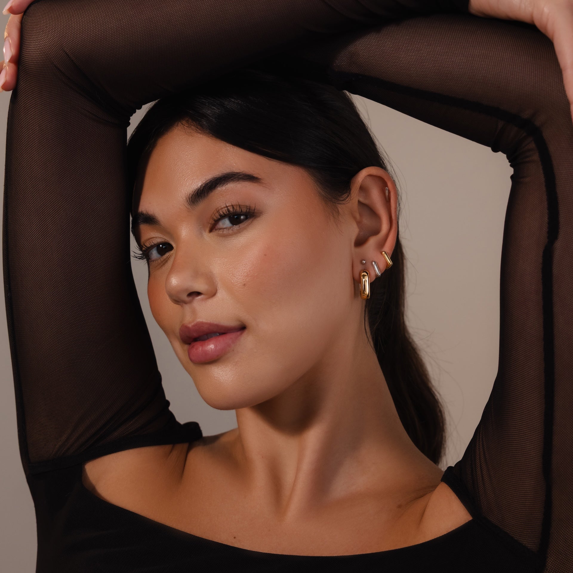 A woman with long dark hair poses with her arm raised, wearing a black mesh top and Bold Link Hoops, giving her look a modern edge with these statement earrings.