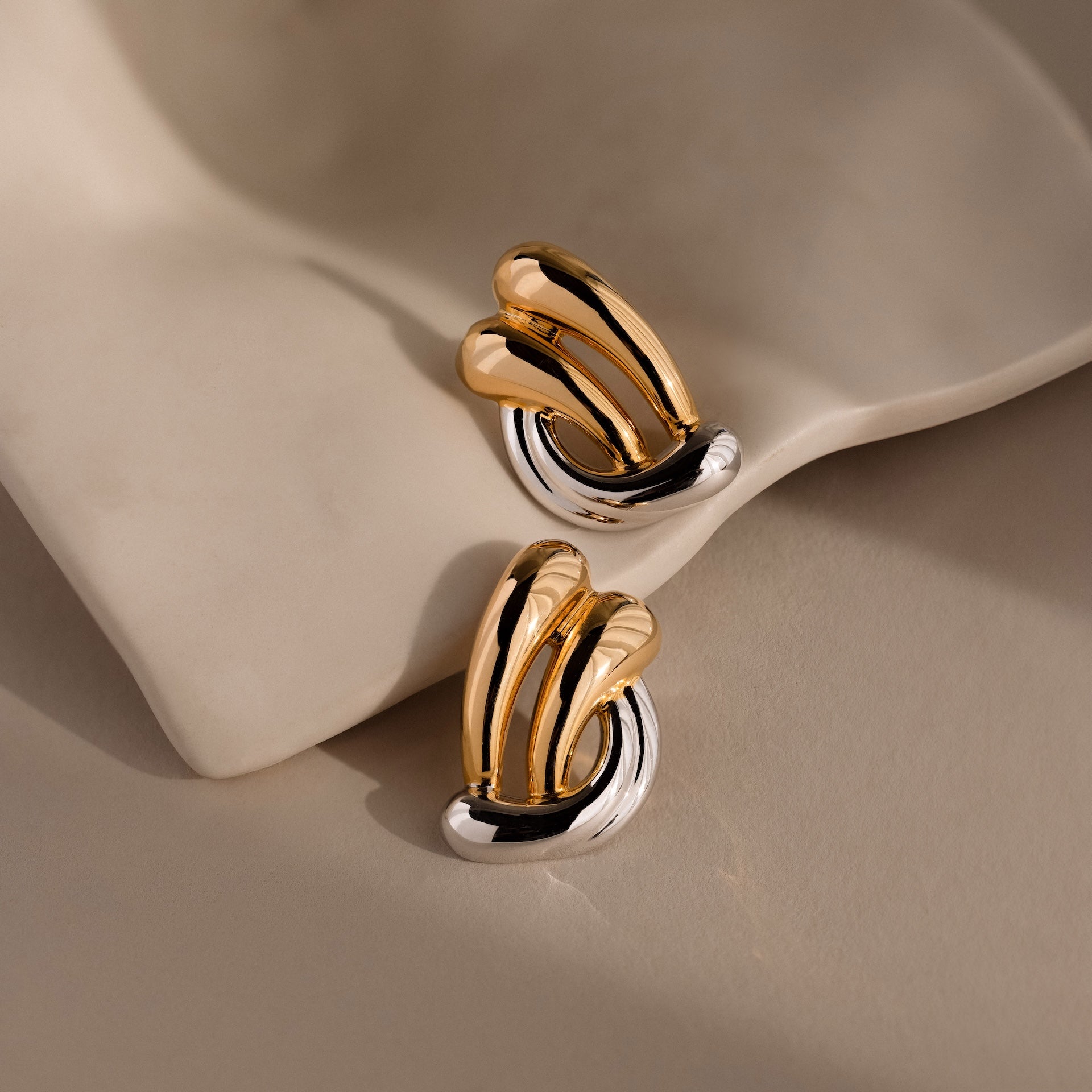 The Mixed Metal Raindrop Studs feature an abstract knot design in gold and silver, shown on a soft beige fabric background.