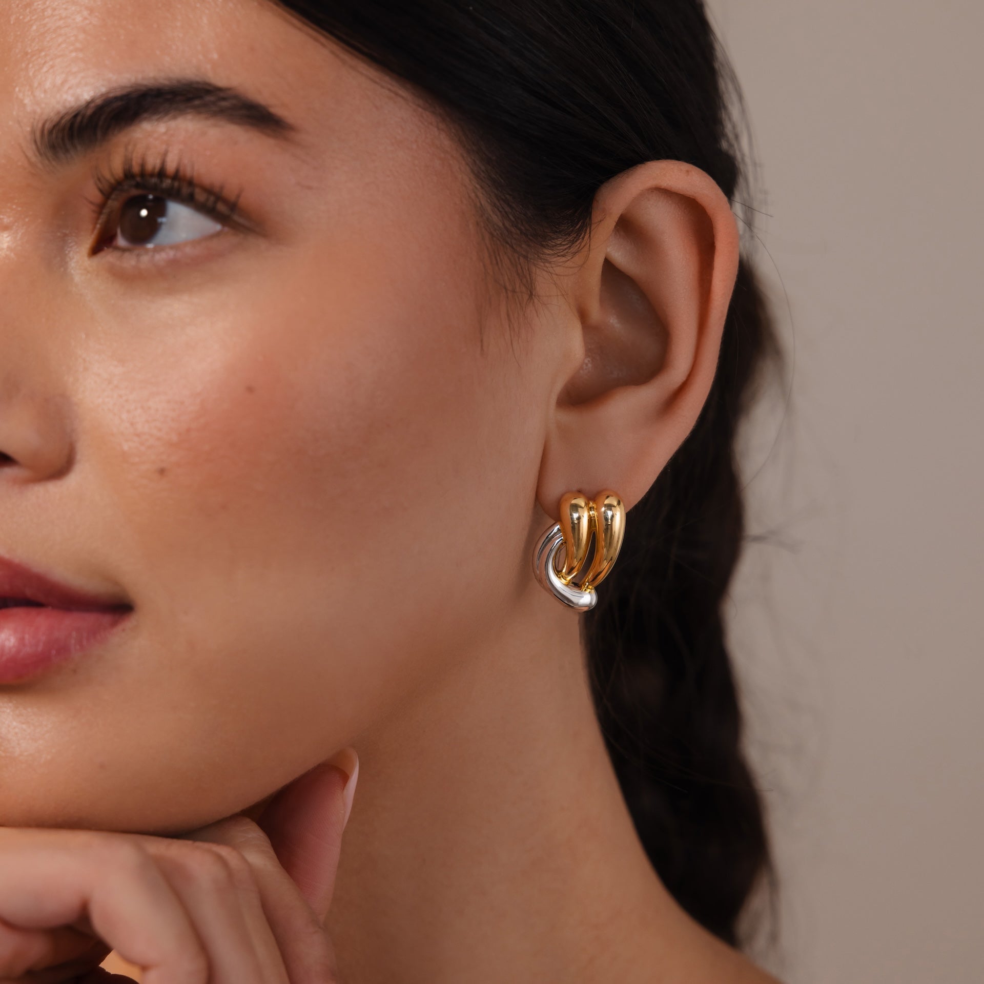 A woman wears our Mixed Metal Raindrop Studs—sculptural gold and silver earrings—while gently touching her chin, embodying an elegant look.