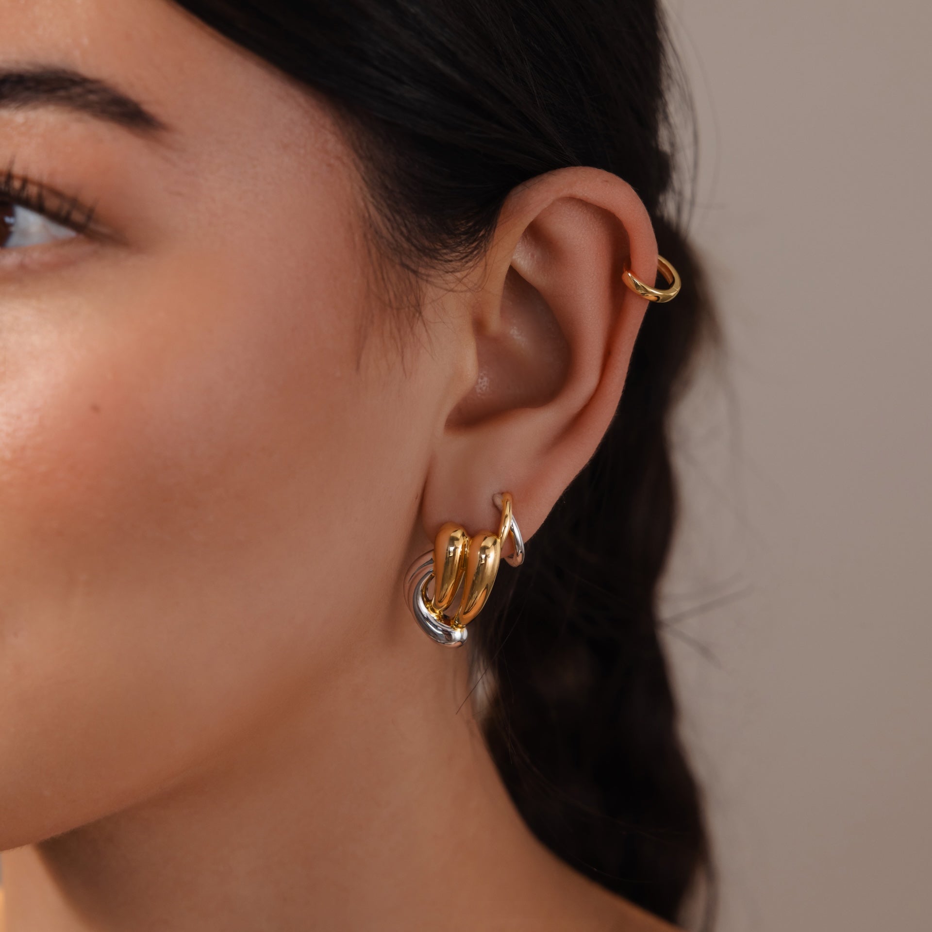 Close-up of a woman's ear adorned with the Mixed Metal Raindrop Studs: sculptural gold and silver earrings styled as three hoops and a gold cuff on the upper ear.