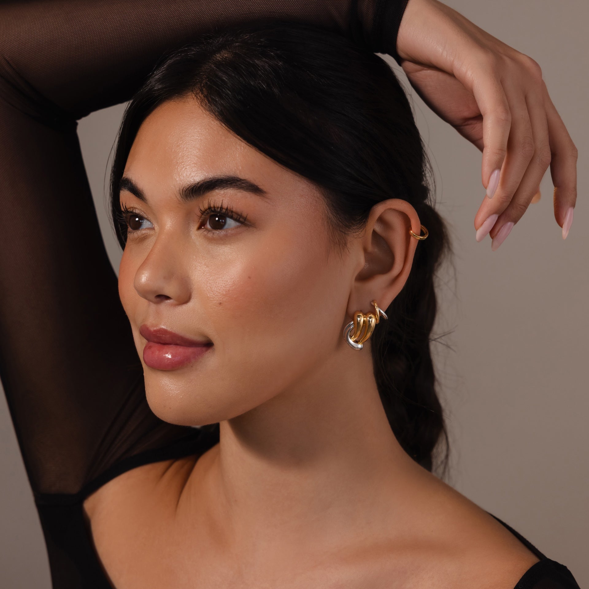 Woman with long dark hair poses in a sheer black top and raises her hand over her head, showcasing Mixed Metal Raindrop Studs—sculptural gold and silver earrings.