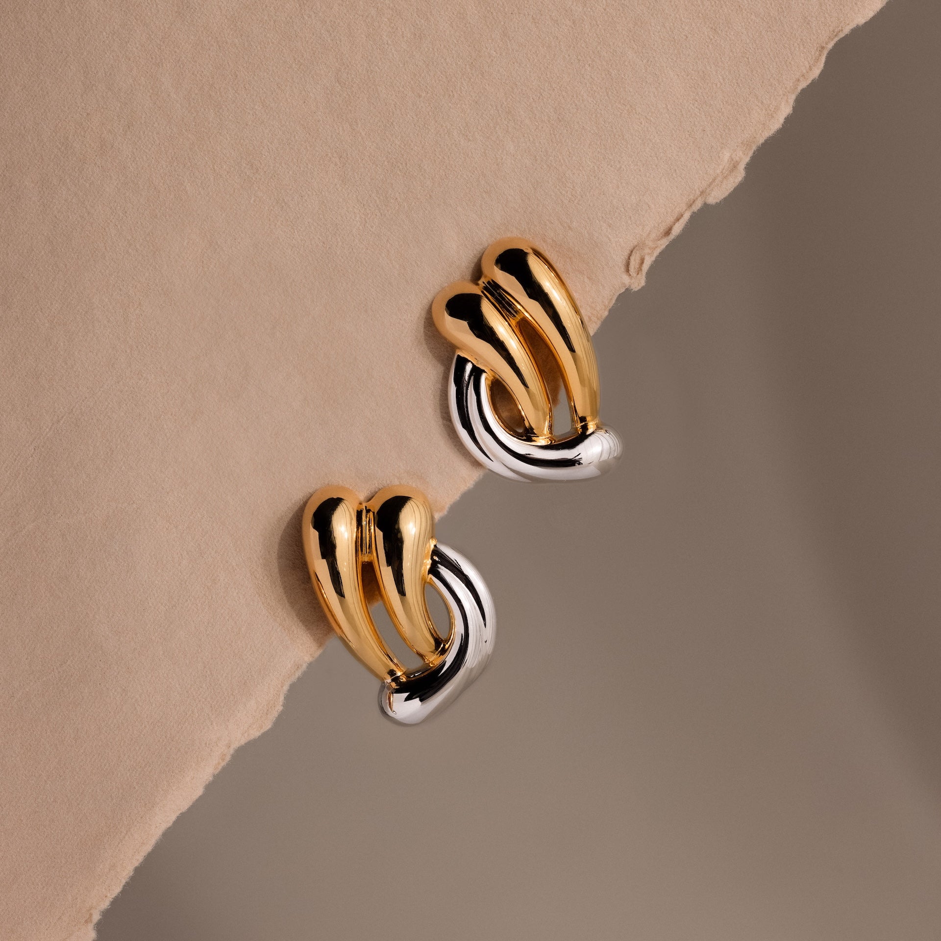 Mixed Metal Raindrop Studs with a sculptural chunky hoop design, crafted in gold and silver, displayed on a beige textured surface.