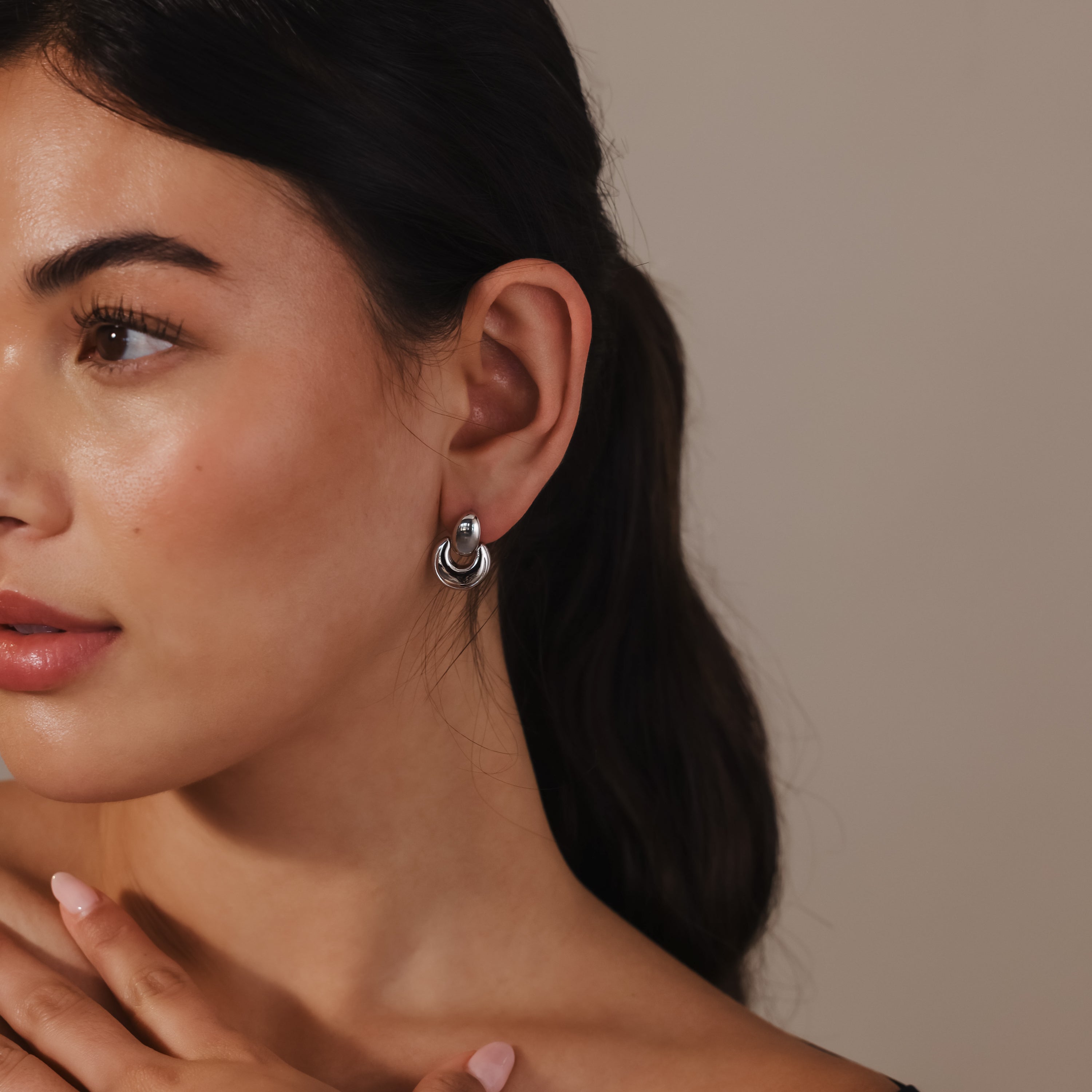 A woman with her hand on her chest showcases the Eclipse Dome Studs, highlighting the allure of modern sculptural earrings.