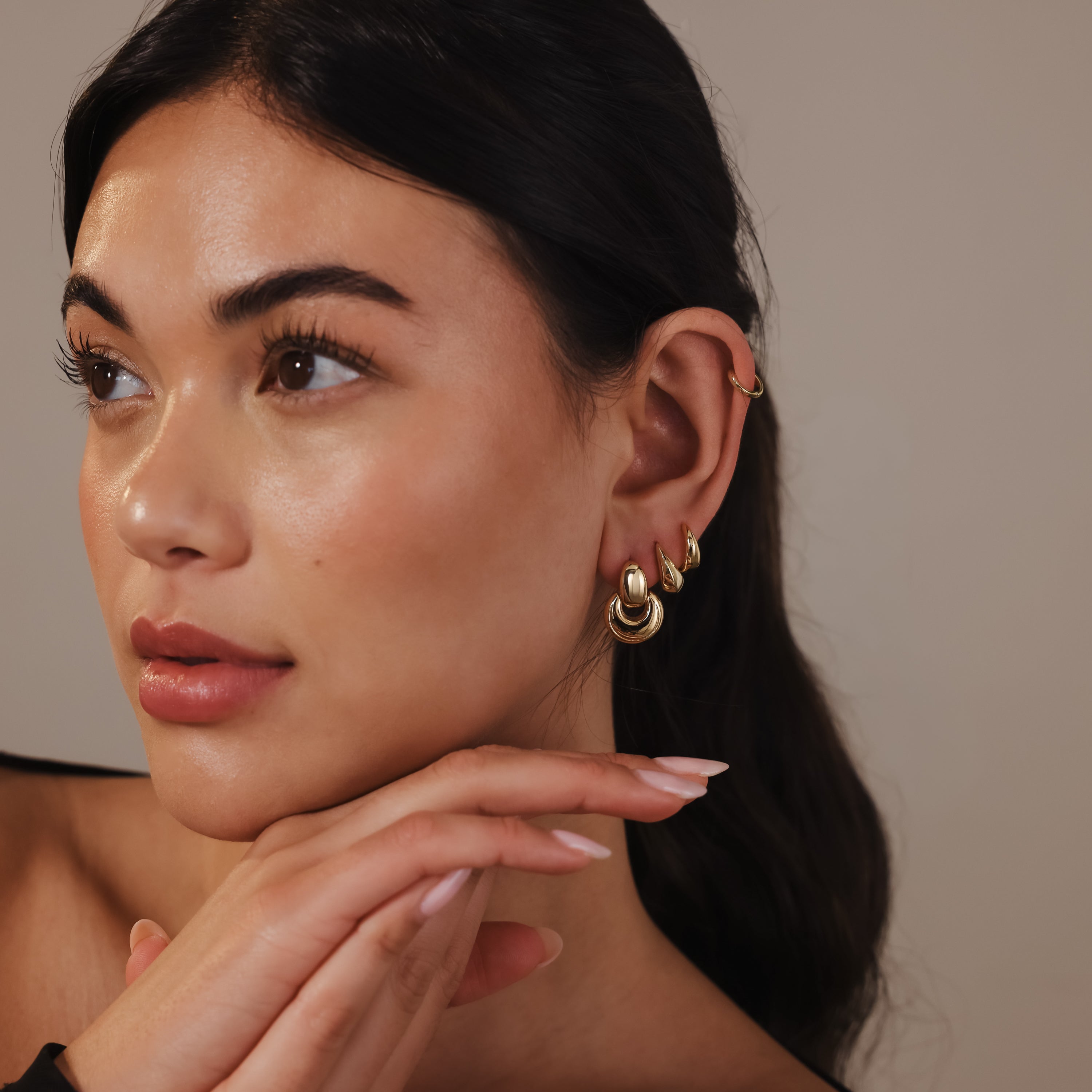 A woman with glowing skin wears Eclipse Dome Studs in 18K Gold, resting her chin on her hands and looking to the side—an elegant touch of modern jewelry.