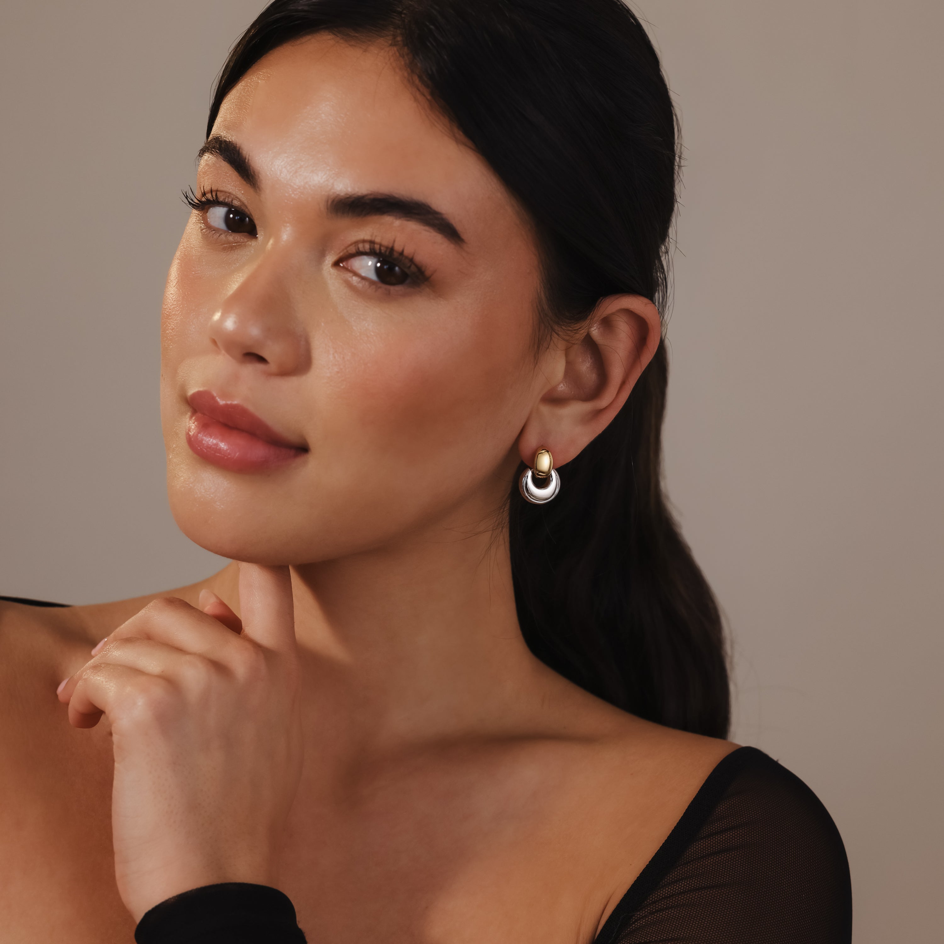 A woman with long dark hair wearing Eclipse Dome Studs in Mixed Metal, touching her chin and posing in a black top—an elegant showcase of modern jewelry.