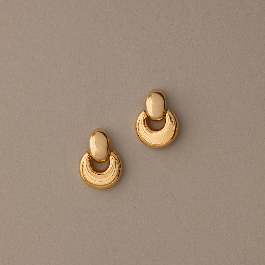 The Eclipse Dome Studs in 18K Gold are semi-circular, shiny dome earrings shown on a plain taupe background—perfect for fans of modern jewelry.