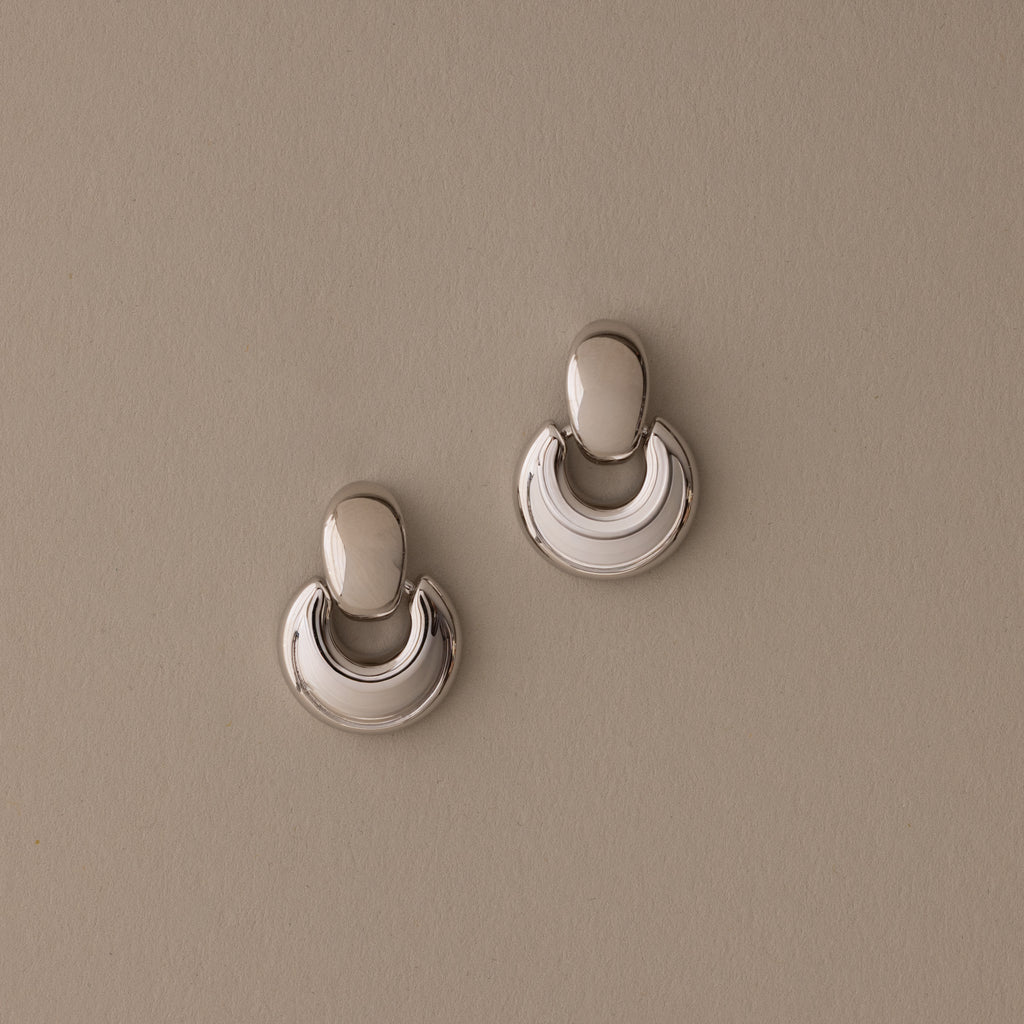 A pair of Eclipse Dome Studs in White Gold, featuring a shiny, crescent-shaped sculptural design, rests on a plain beige background.