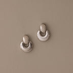 A pair of Eclipse Dome Studs in White Gold, featuring a shiny, crescent-shaped sculptural design, rests on a plain beige background.