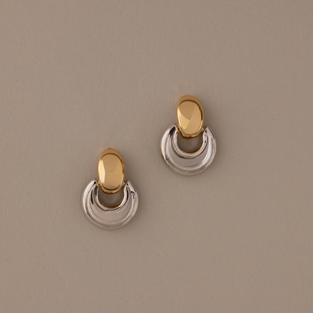The Eclipse Dome Studs in Mixed Metal feature oval tops and curved half-moon bottoms, highlighting sculptural design against a beige background.