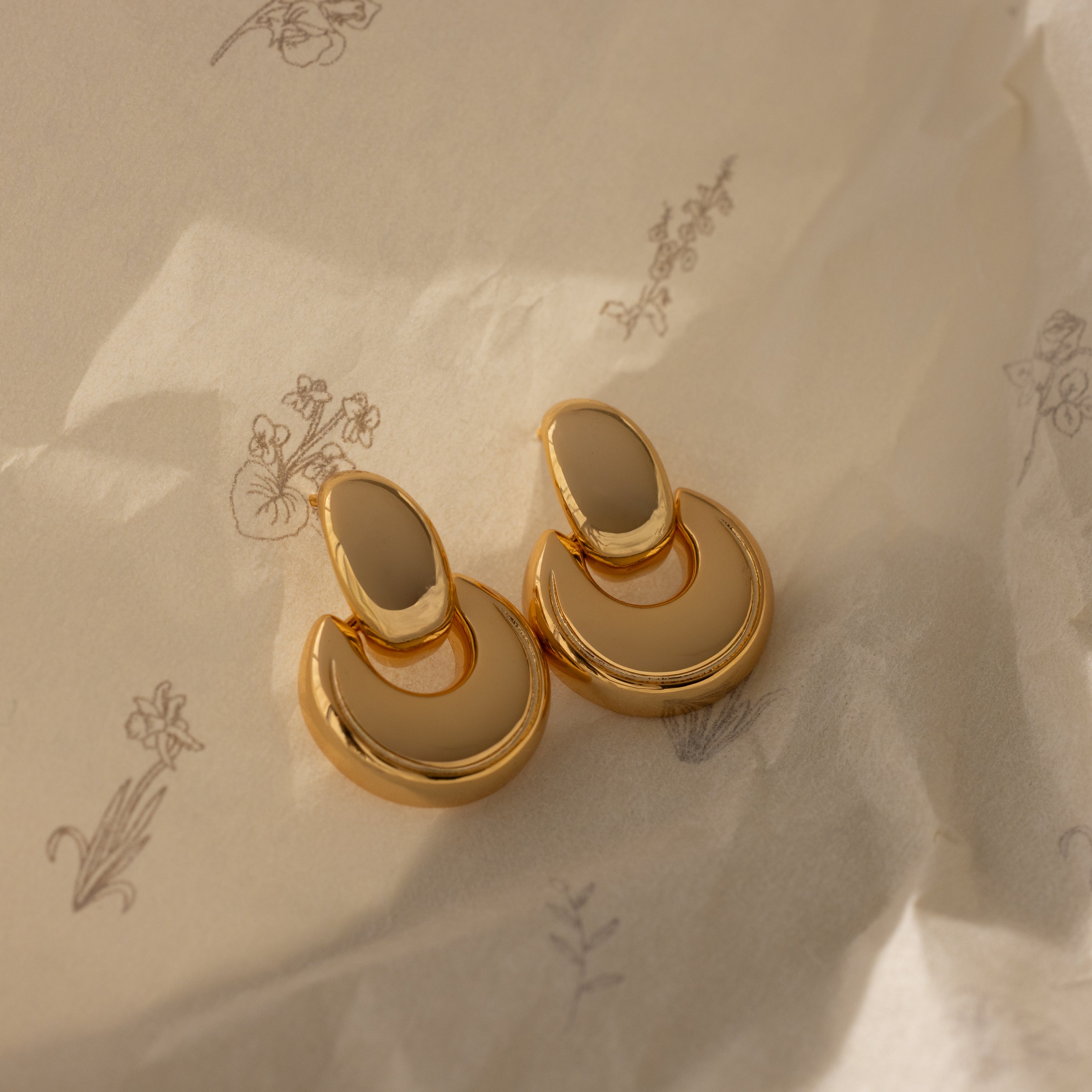 The Eclipse Dome Studs in 18K Gold, with their gold and beige crescent shape, rest on crumpled paper featuring delicate floral sketches, capturing the essence of modern jewelry.