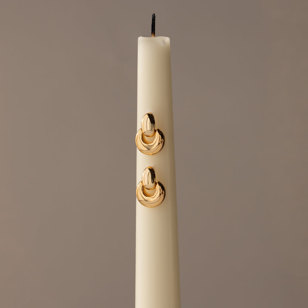 A white candle displays sculptural earrings and Eclipse Dome Studs in 18K gold, set against a neutral background for a modern jewelry showcase.