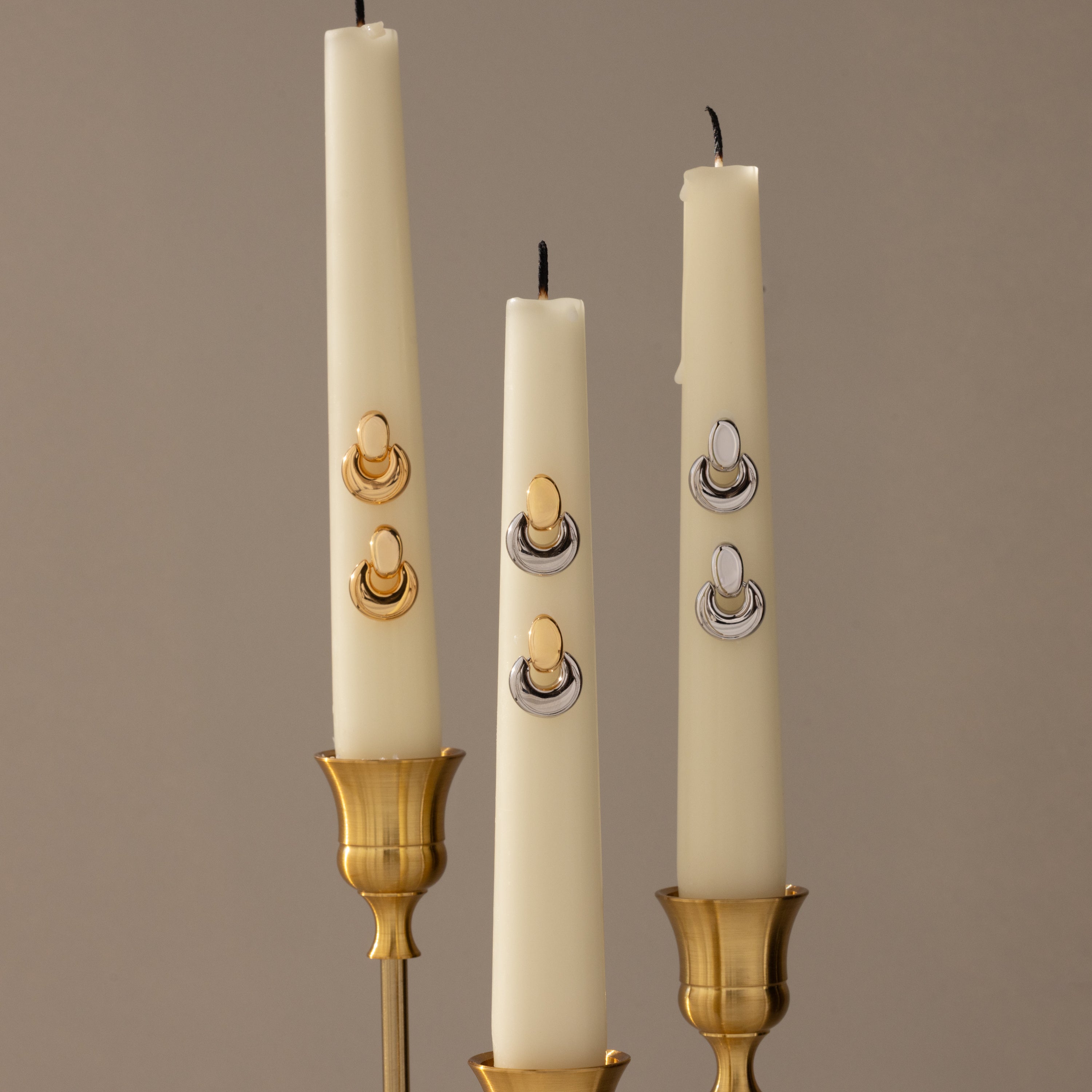 Three white candles in brass holders, each adorned with sculptural earrings like the Eclipse Dome Studs in Mixed Metal, create a striking display of modern jewelry against a neutral background.