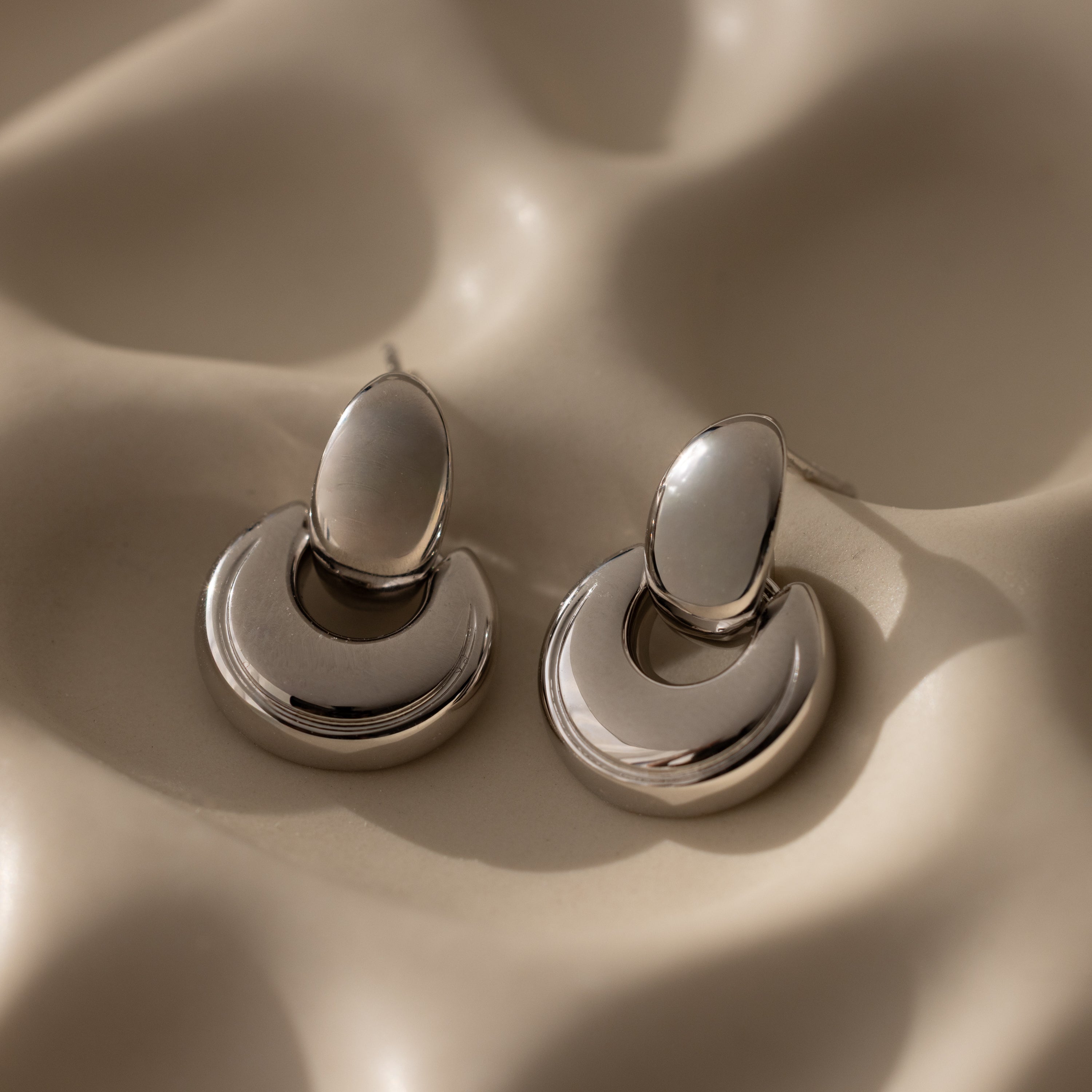 The Eclipse Dome Studs, with their silver crescent design, rest on a wavy beige surface with soft shadows, capturing the essence of modern jewelry elegance.