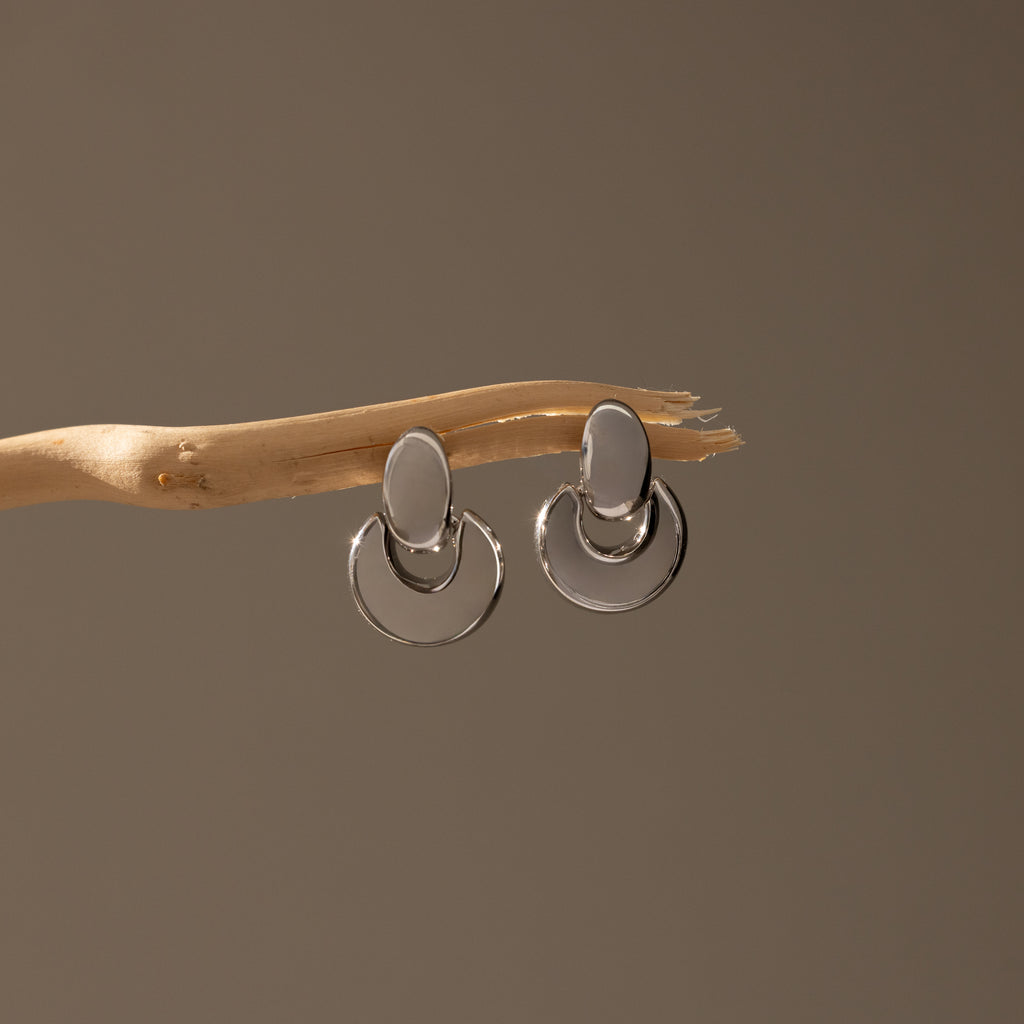 Eclipse Dome Studs in White Gold, featuring sculptural oval and circular forms, hang on a slender, light wooden branch against a neutral background, highlighting the refined elegance of modern jewelry design.