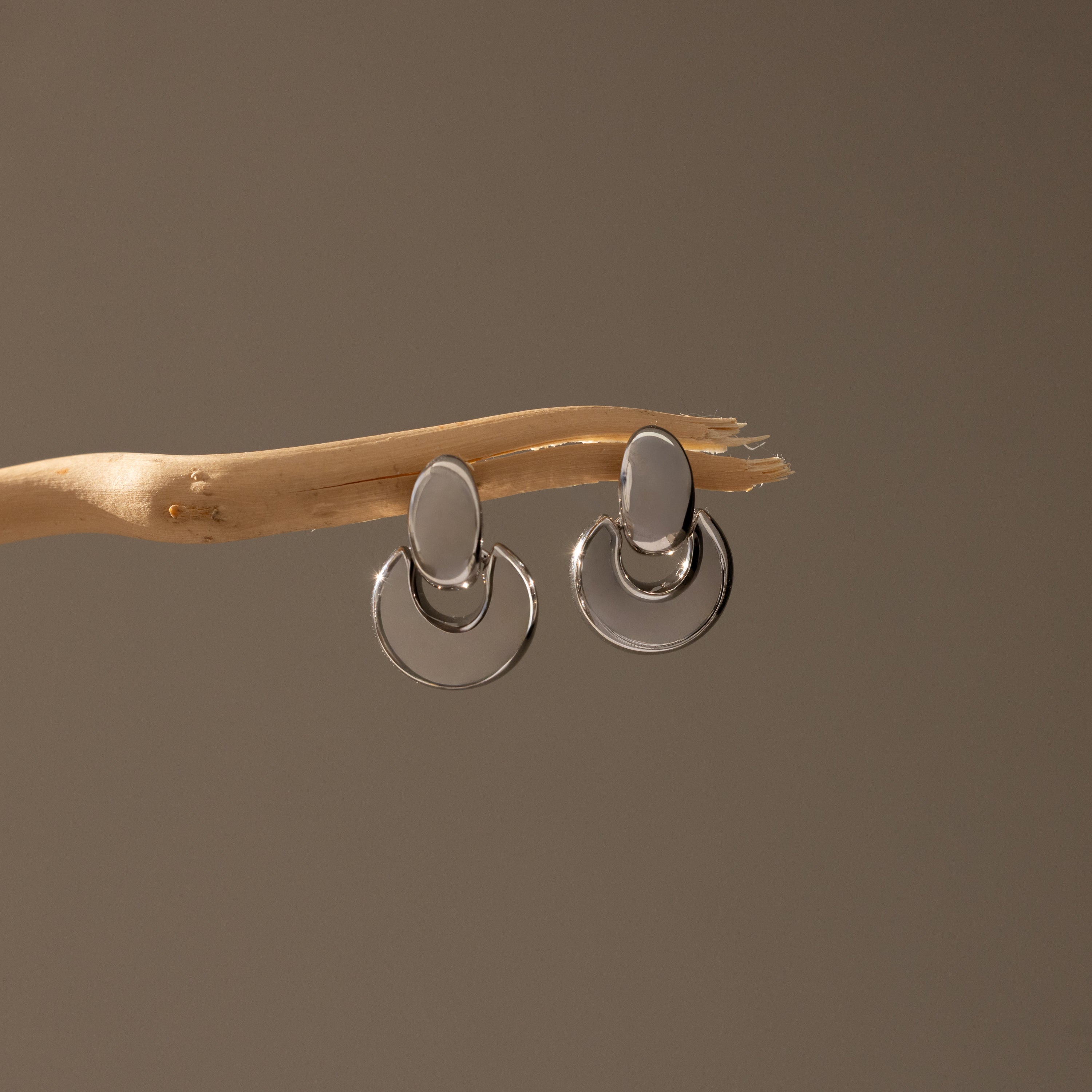 Eclipse Dome Studs in White Gold, featuring sculptural oval and circular forms, hang on a slender, light wooden branch against a neutral background, highlighting the refined elegance of modern jewelry design.