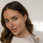 Woman with wavy brown hair and a light-colored top smiles softly at the camera, showcasing the Dainty Pearl Studs in 18K Gold.