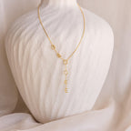 The Lariat Name Necklace features gold dangling letters spelling “BROOKE,” displayed on a white textured bust—a stunning personalized jewelry piece perfect for everyday wear.