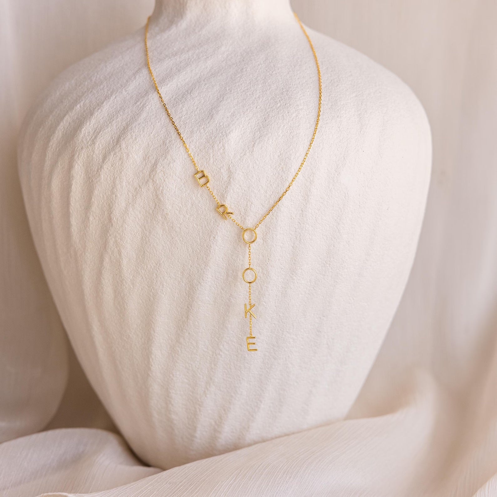 The Lariat Name Necklace features gold dangling letters spelling “BROOKE,” displayed on a white textured bust—a stunning personalized jewelry piece perfect for everyday wear.