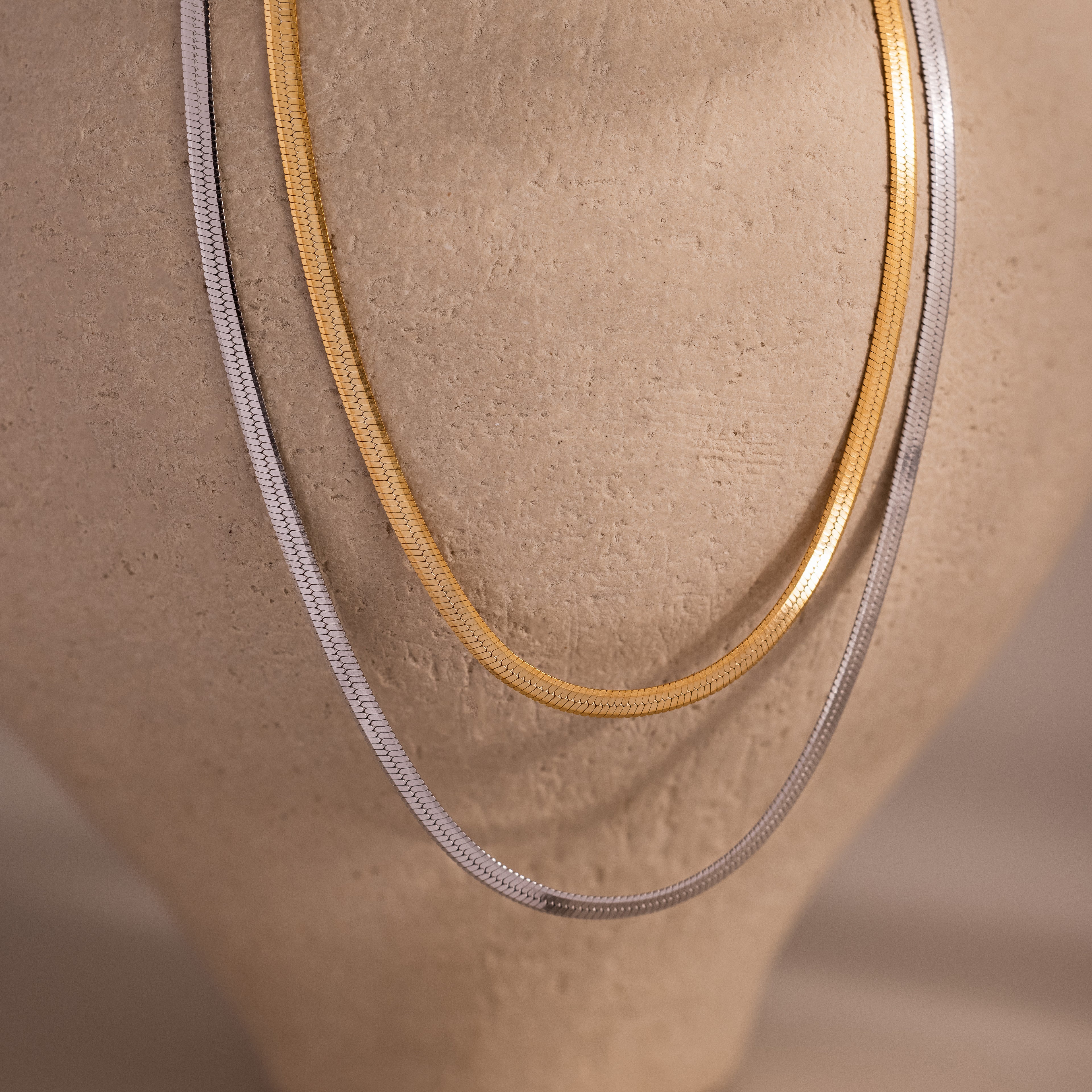 Two herringbone chain necklaces, one gold and one silver, draped over a mannequin bust.