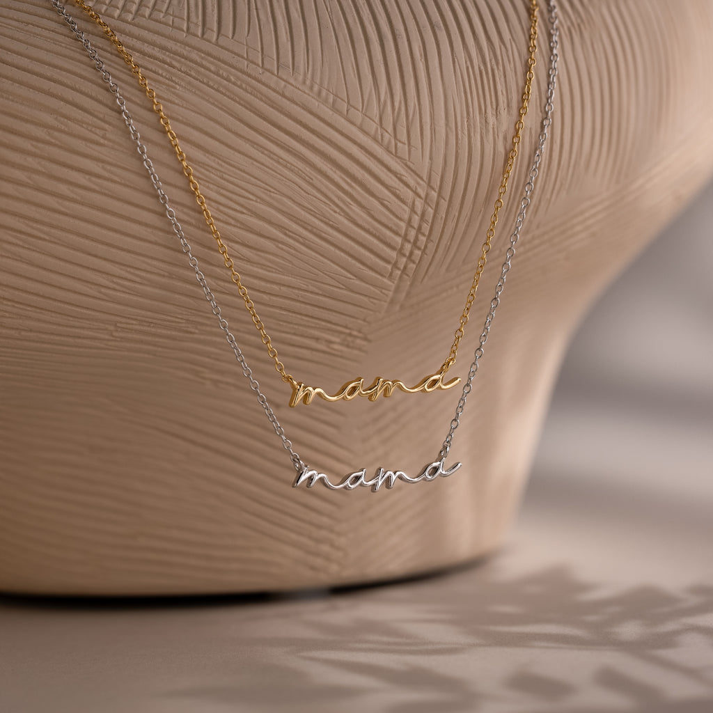 Two 'mama' pendant necklaces in gold and silver on a textured beige background