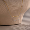 Two 'mama' pendant necklaces in gold and silver on a textured beige background