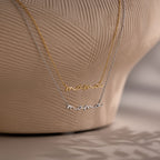 Two 'mama' pendant necklaces in gold and silver on a textured beige background