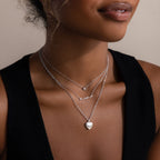 Woman wearing multiple silver necklaces and one with a heart pendant on a neutral background