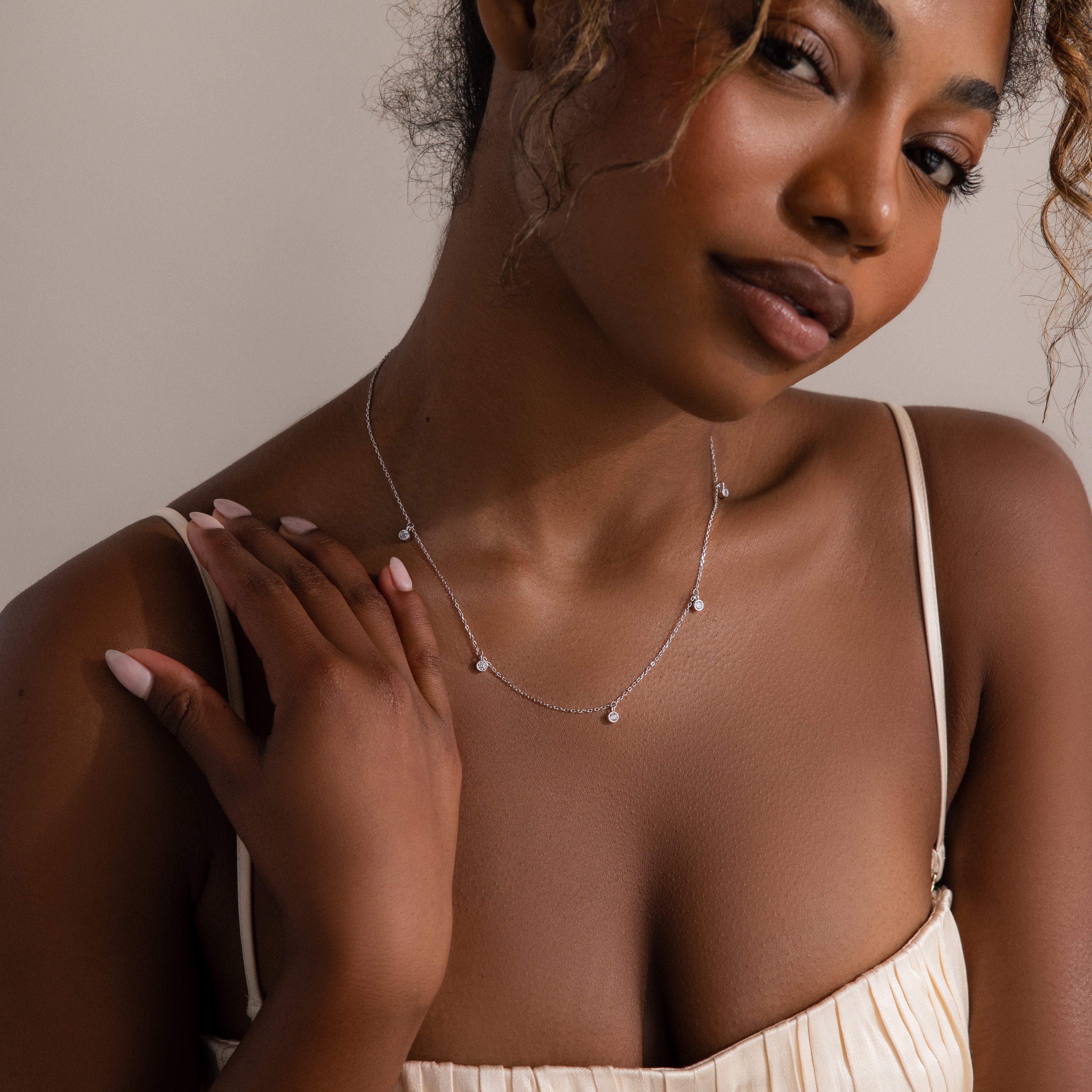 A woman in a cream dress touches her collarbone and gazes at the camera, showcasing the Diamond Station Necklace—a modern yet timeless accessory.