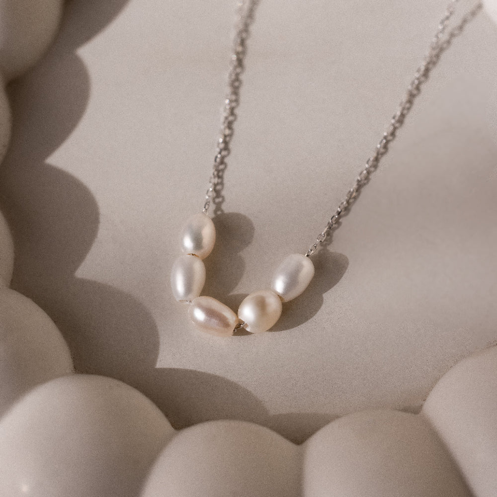 The Pearl Bead Necklace in Silver features five white pearls on a delicate silver chain, displayed on a soft beige surface with rounded shapes. Ideal as a bridesmaids or Mothers Day gift.