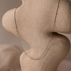 The Satellite Necklace in Silver drapes over a textured, abstract beige sculpture—an ideal layering piece for effortless elegance.