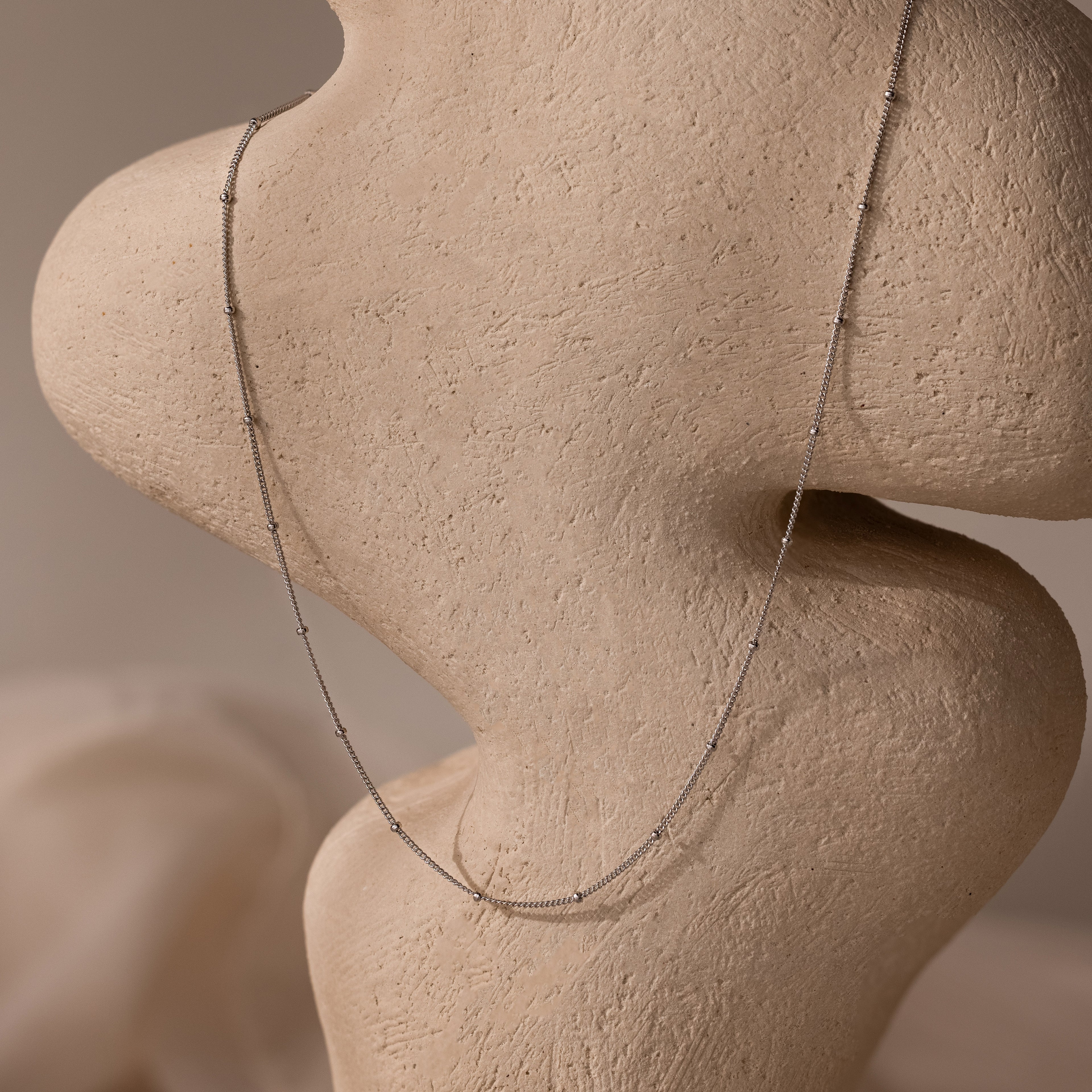 The Satellite Necklace in Silver drapes over a textured, abstract beige sculpture—an ideal layering piece for effortless elegance.