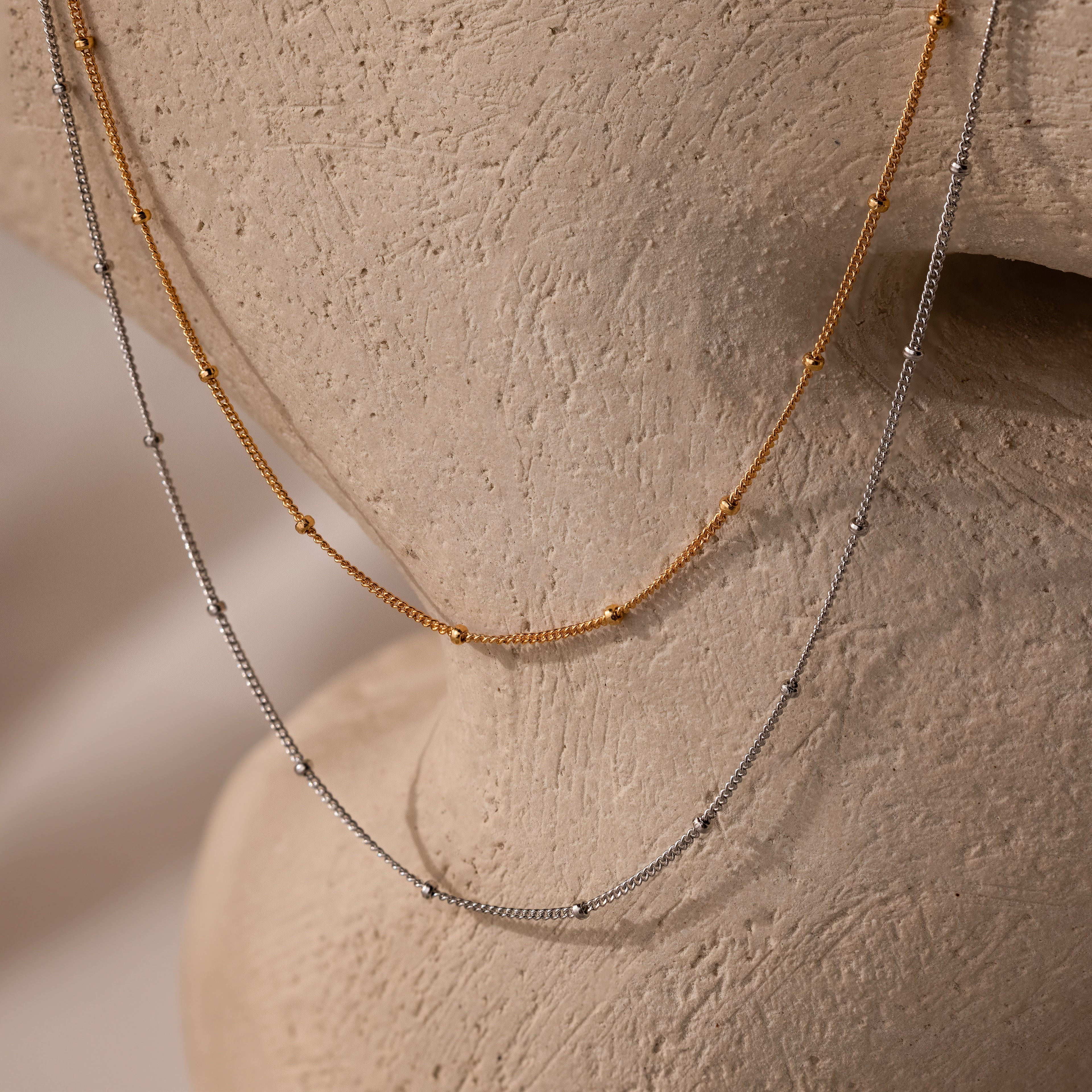 These Satellite Necklaces, a simple gold and silver beaded chain perfect for layering, is draped over a textured beige sculpture.
